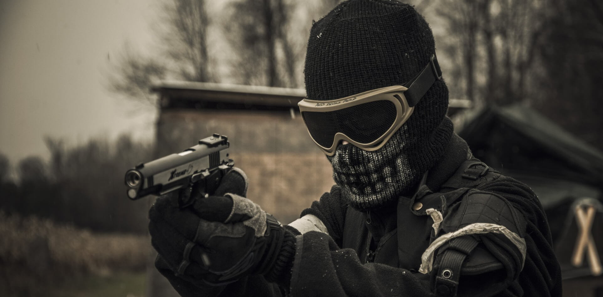 hombre con una pistola apuntando hacia la izquierda posando para la foto con su vestimenta con predominancia negra con las gafas de airsoft de proteccion