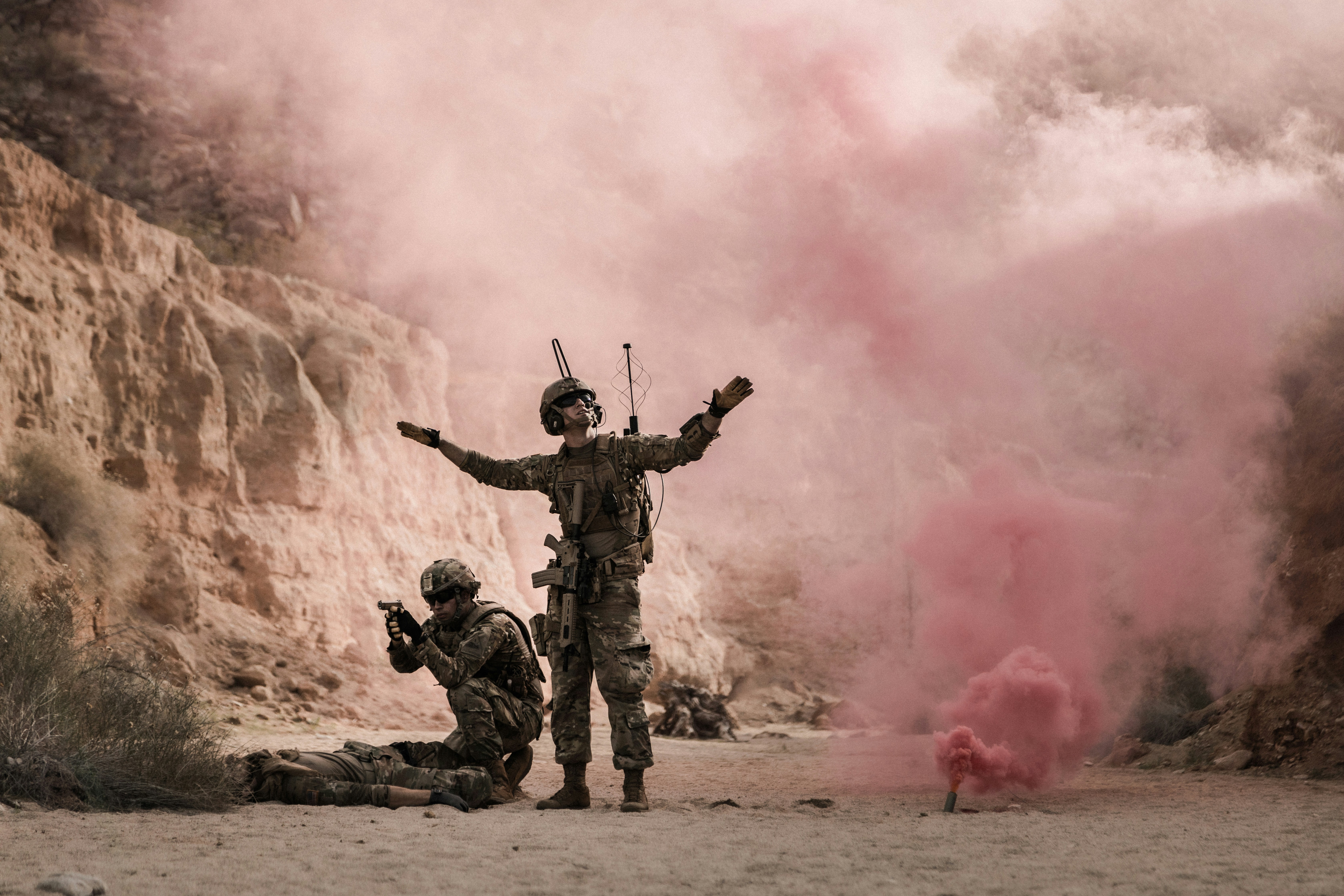 tres militares en un terreno arido con humo rojo detras de fondo , simulando una extraccion de un hombre caido