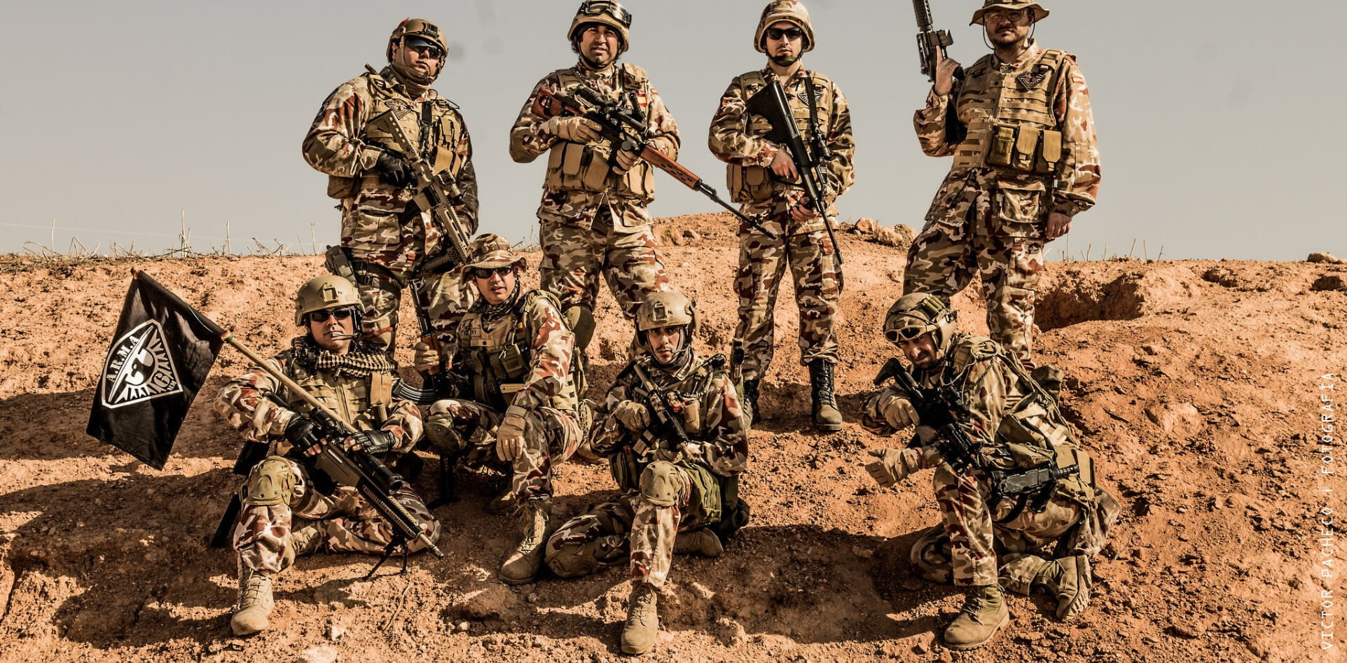 un grupo de militares posando para la foto con su vestimenta adecuada, con su campo arido de fondo y una bandera que representa su organizacion